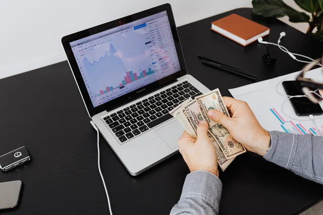 Person counting dollar bills with financial graphs on a laptop screen, symbolizing financial analysis and planning. SAAS marketing hidden costs often arrive after the contract is signed